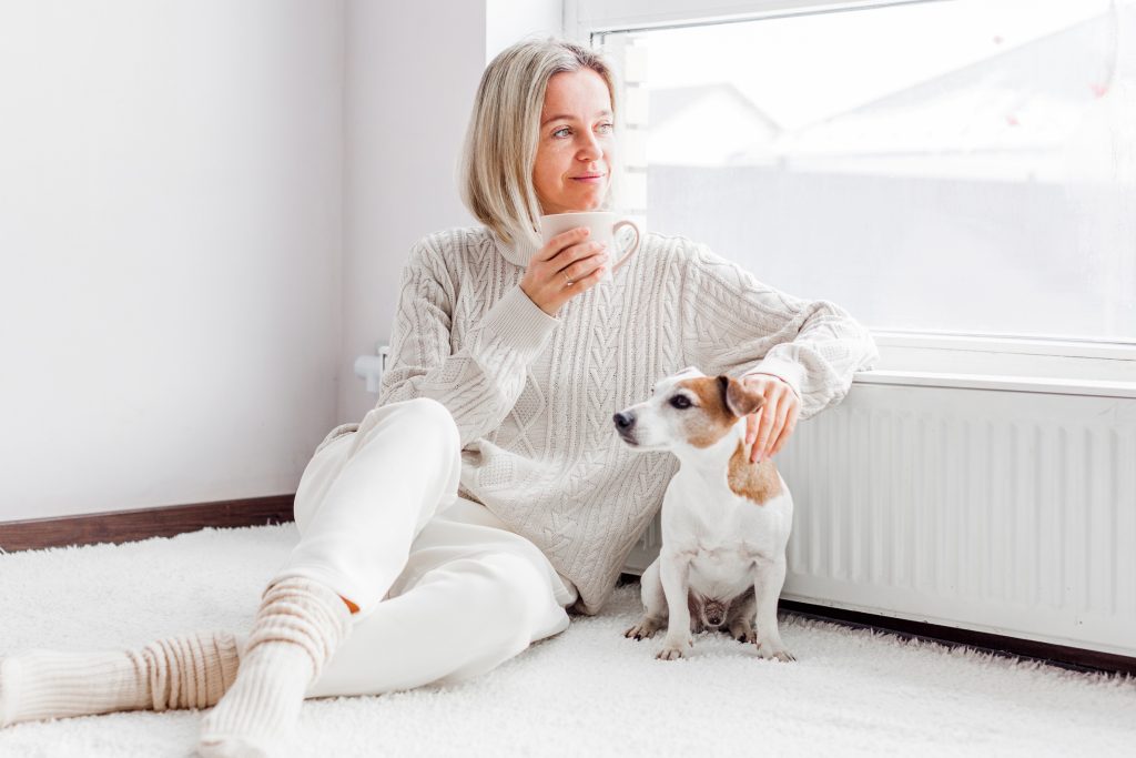Nainen istuu koiran kanssa patterin ja ikkunan vieressä.